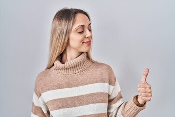 Young blonde woman wearing turtleneck sweater over isolated background looking proud, smiling doing thumbs up gesture to the side