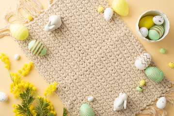 Easter celebration concept. Top view photo of bowl with colorful easter eggs ceramic bunnies cloth napkin and yellow mimosa flowers on isolated beige background