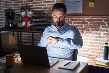 Hispanic man with beard working at the office at night checking the time on wrist watch, relaxed...