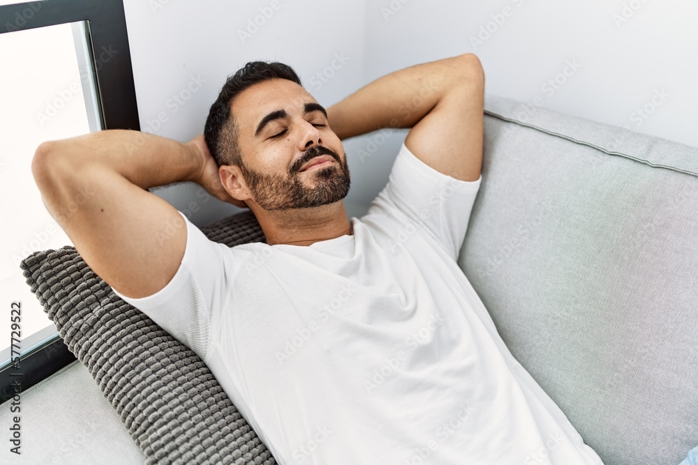 Sticker Young hispanic man relaxed with hands on head lying on sofa at home