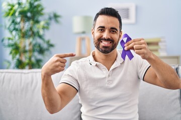 Young hispanic man with beard holding purple ribbon awareness pointing finger to one self smiling...