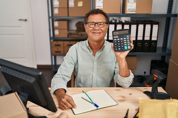 Senior man working at small business ecommerce with calculator looking positive and happy standing and smiling with a confident smile showing teeth