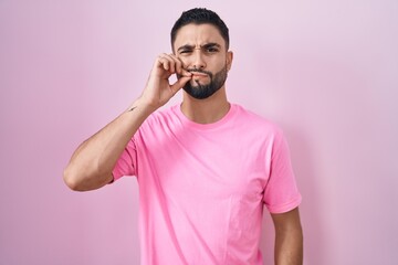 Hispanic young man standing over pink background mouth and lips shut as zip with fingers. secret and silent, taboo talking