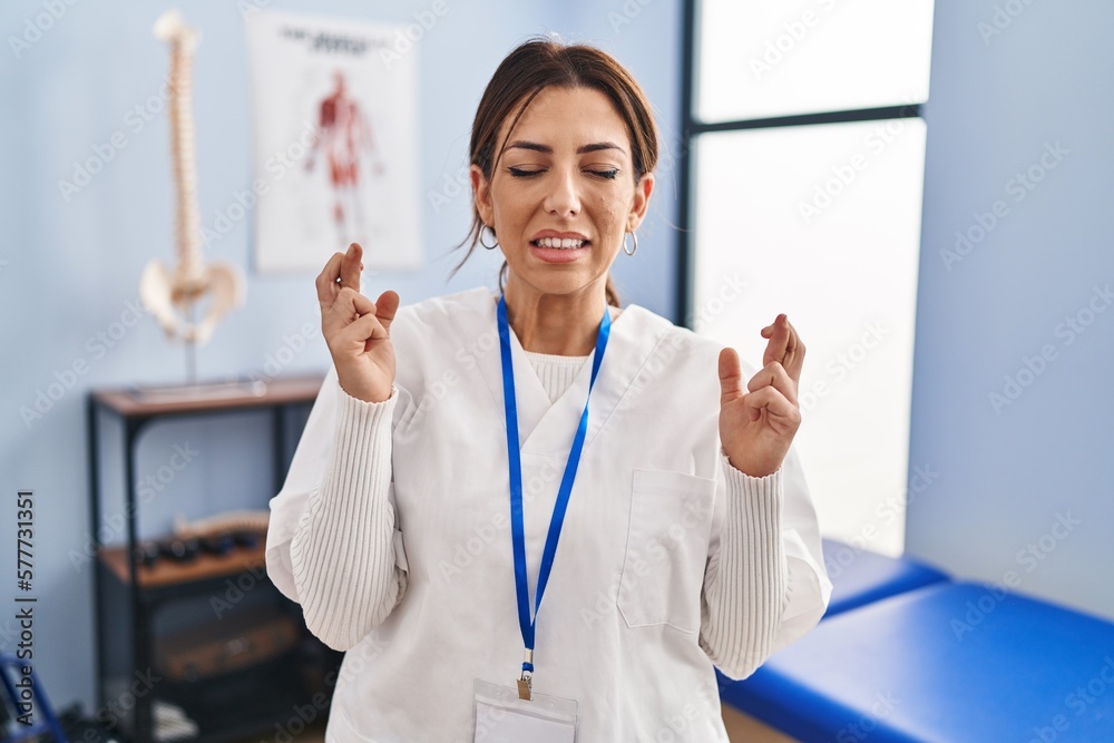 Canvas Prints Young brunette woman working at pain recovery clinic gesturing finger crossed smiling with hope and eyes closed. luck and superstitious concept.