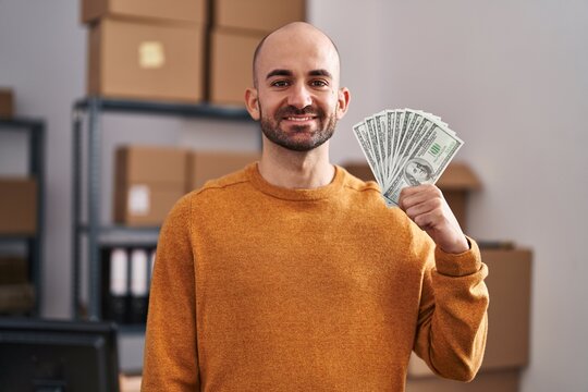 Young Bald Man With Beard Working At Small Business Ecommerce Holding Money Looking Positive And Happy Standing And Smiling With A Confident Smile Showing Teeth