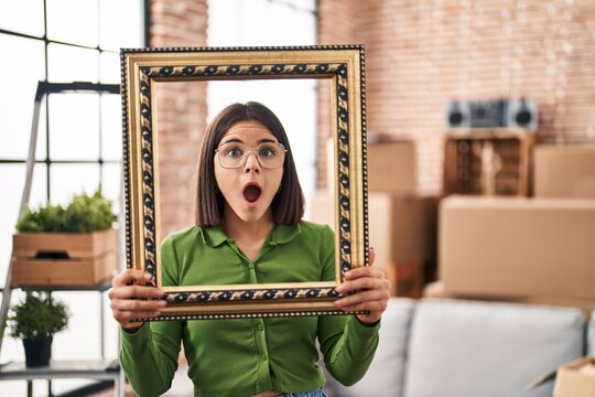 Young hispanic woman at new home holding empty frame afraid and shocked with surprise and amazed expression, fear and excited face.