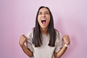 Young hispanic woman standing over pink background angry and mad screaming frustrated and furious, shouting with anger. rage and aggressive concept.