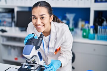 Young hispanic woman scientist using microscope at laboratory - obrazy, fototapety, plakaty