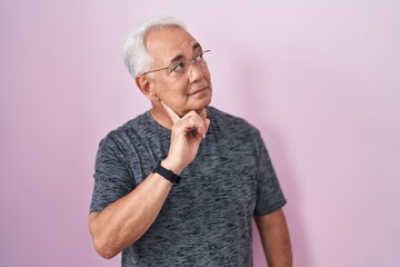 Middle age man with grey hair standing over pink background with hand on chin thinking about question, pensive expression. smiling with thoughtful face. doubt concept.