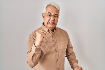 Hispanic senior man wearing glasses angry and mad raising fist frustrated and furious while shouting with anger. rage and aggressive concept.