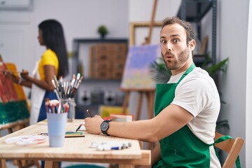 Hispanic man at painter studio scared and amazed with open mouth for surprise, disbelief face