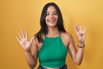 Brunette woman standing over yellow background showing and pointing up with fingers number nine...