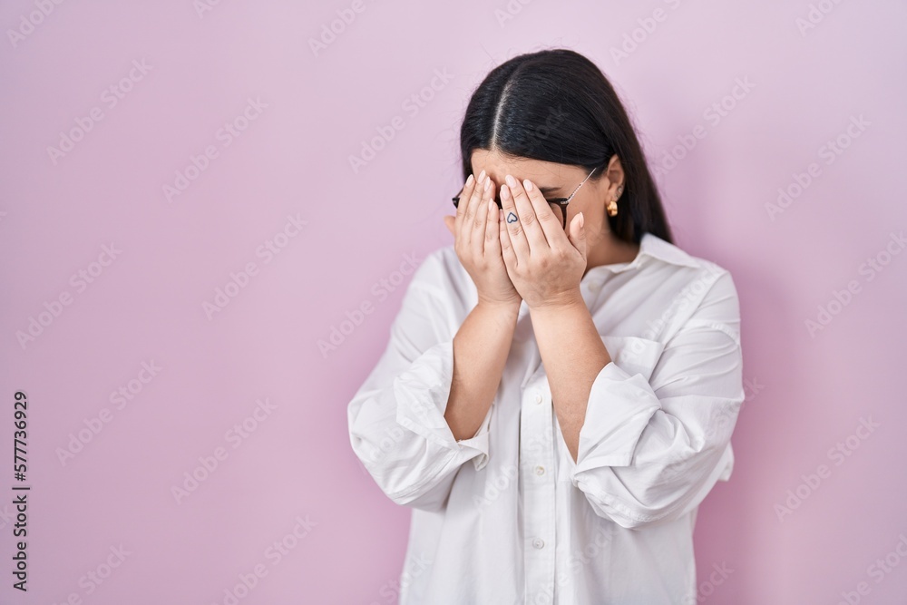 Poster young brunette woman standing over pink background with sad expression covering face with hands whil