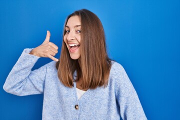 Young woman standing over blue background smiling doing phone gesture with hand and fingers like talking on the telephone. communicating concepts.