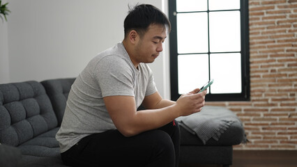 Young chinese man using smartphone sitting on sofa at home