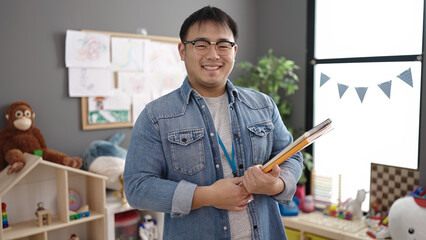 Young chinese man teacher smiling confident holding books at kindergarten