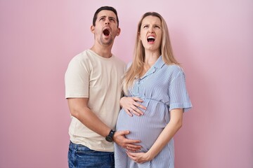 Young couple expecting a baby standing over pink background angry and mad screaming frustrated and...
