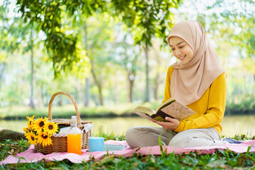 Happy cheerful Asian beautiful muslim woman sitting on the ground at the park and drinking a cup of...