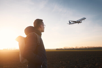 Man with backpack looking up to airplane landing at airport during beautiful sunset. .