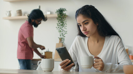 Ethnic multiracial couple on kitchen Arabian woman sit at table with mobile phone browsing internet work freelance at home use smartphone. Husband man help wife with cooking make coffee on background 