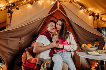 Happy lovely couple relaxing in glamping on summer evening and drinking wine near cozy bonfire. Luxury camping tent for outdoor recreation and recreation. Lifestyle concept