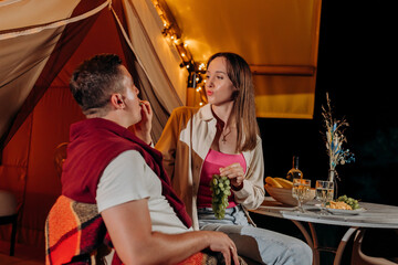 Happy lovely couple relaxing in glamping on summer evening and eating grapes near cozy bonfire. Luxury camping tent for outdoor recreation and recreation. Lifestyle concept