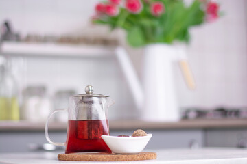 a glass teapot with tea and dessert stands on a white kitchen table against the background of a kitchen interior