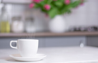 a white cup of coffee stands on a white kitchen table against the background of a kitchen interior Close up