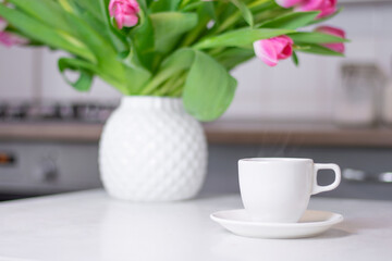 a white cup of coffee and vase of flowers stands on a white kitchen table against the background of a kitchen interior