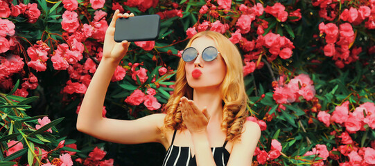 Portrait of beautiful woman taking selfie by smartphone blowing her lips with red lipstick sending sweet air kiss in spring garden on pink flowers roses background