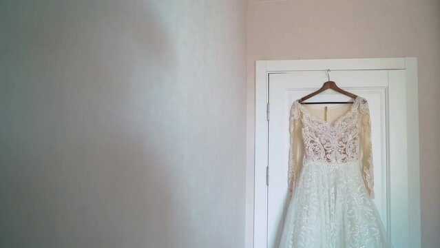 A beautiful white wedding dress hangs on a hanger on the door in the bedroom. Morning of the bride, wedding day. Women's fashionable clothes for the holiday.