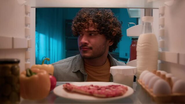 View inside from refrigerator Indian sly man with curly hair open fridge at night evening in kitchen hungry guy male looking around take out slice of pizza overeating at nighttime unhealthy nutrition