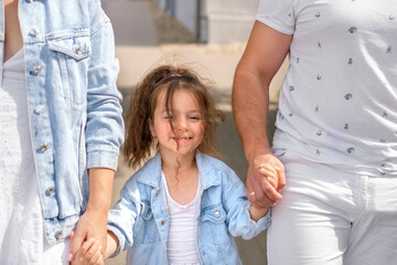 A young family leads a little curly-haired daughter by the hand along a city street on a sunny summer day. A proud child under the protection of parents