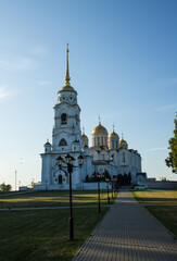 Beautiful landscape - the white-stone Assumption Cathedral with a bell tower in Vladimir Russia on a sunny summer morning among the green foliage of trees against a blue cloudless sky