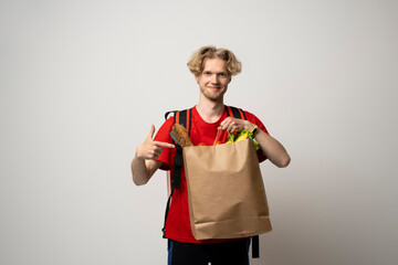 Food delivery service. Portrait of pleased delivery man in red uniform smiling while carrying paper bag with food products isolated over white background.