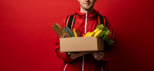 Grocery delivery courier in red uniform with a paper box with food showing a thumb up. Thumb up gesture. Food delivery service concept. - obrazy, fototapety, plakaty