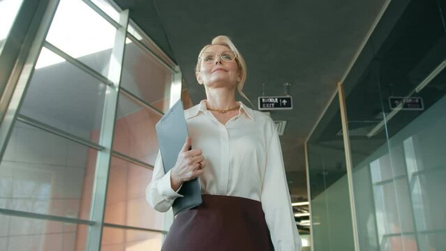 Portrait of focused businesswoman in eyeglasses walking office corridor. Stylish mature confident woman 50s walking in corridor. Serious business woman going with folder in business center hallway 4K