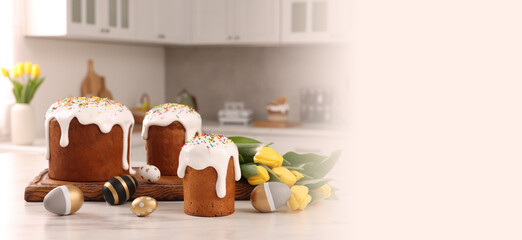 Delicious Easter cakes with sprinkles, painted eggs and beautiful tulips on white marble table in kitchen, space for text. Banner design