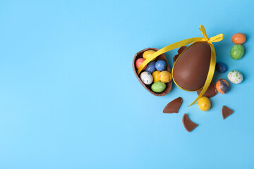 Tasty whole chocolate egg with yellow bow and different candies on light blue background, flat lay. Space for text