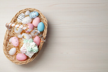 Easter basket with painted eggs and tasty cake on white wooden table, top view. Space for text