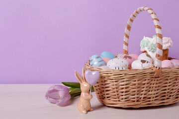 Easter basket with painted eggs, cake, flower and rabbit figure on white wooden table. Space for text