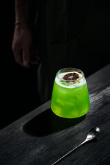 Special green cocktail of mixology, lemon rum, garnished with dried fruit, on black bar table, bartender in the background