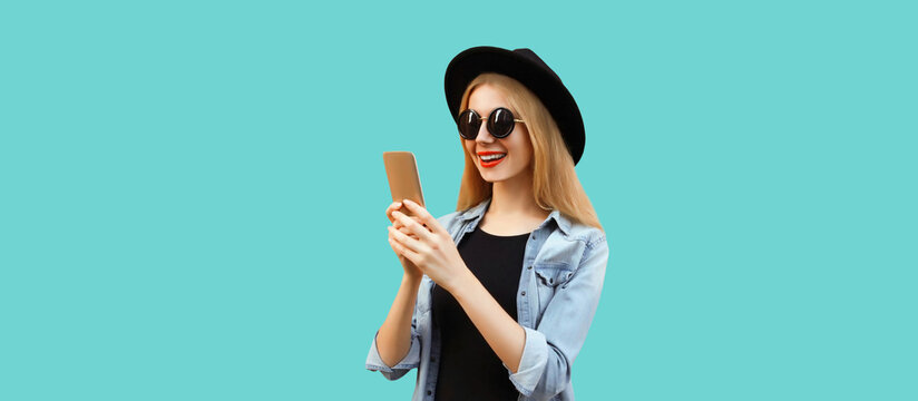 Portrait of happy smiling young woman with smartphone wearing black round hat isolated on blue background