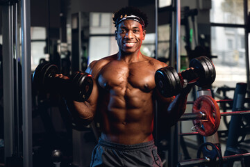 Fototapeta 20s Black and muscular man in a gym showing dumbbells and his muscles. obraz