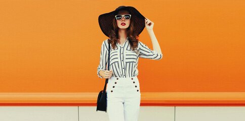 Portrait of beautiful woman model wearing black round straw summer hat, handbag and white striped...
