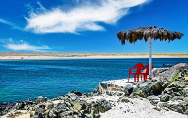 2 lonely empty red chairs under a palm leaf parasol on a stone terrace on a rocky beach on a...