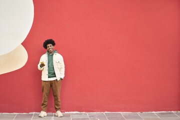 Happy smiling cool generation z young African American ethnic stylish hipster guy model standing at red city urban wall outdoors using cell phone mobile device, looking at camera, holding cellphone. - obrazy, fototapety, plakaty