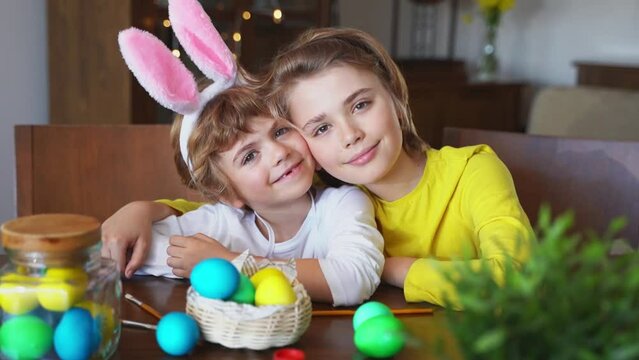 Easter Sunday Family holiday traditions. Two caucasian happy siblings kids with bunny ears posing smiling looking at camera decorated eggs while sitting together at home table. Kids having fun