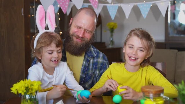 Easter Family traditions. Father and two caucasian happy children with bunny ears playing with Easter decorated eggs while sitting together at home holiday table.