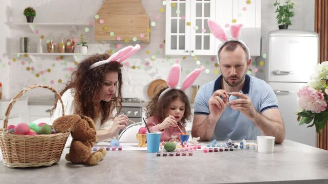 Portrait of loving parents in rabbit ears coloring traditional eggs. Holiday leisure activity with painting and brushing Easter food at family home. High quality 4k footage